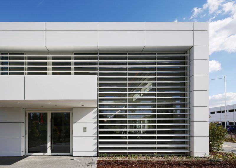 Fassade eines modernen Gebäudes mit großen Glasflächen und horizontalen Lamellen vor einem Fenster neben einer Glastür