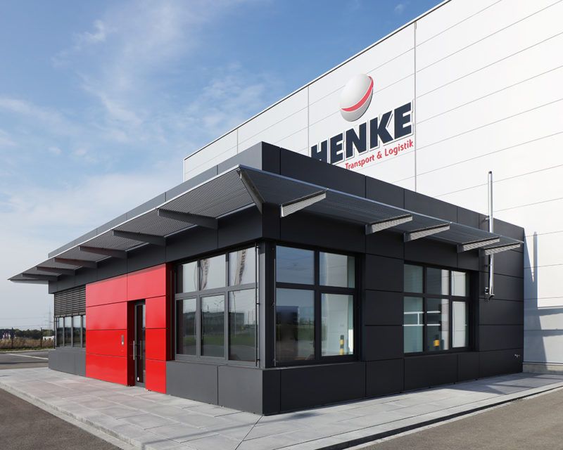 Modernes Firmengebäude mit schwarzer Fassade und roter Eingangstür vor weißer Wand mit Schriftzug 'HENKE Transport & Logistik'