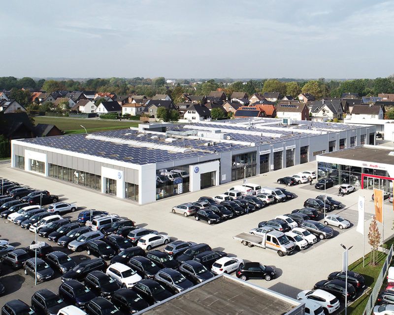 Luftaufnahme eines großen Autohauses mit mehreren Gebäuden, vielen geparkten Autos und einem Wohngebiet im Hintergrund